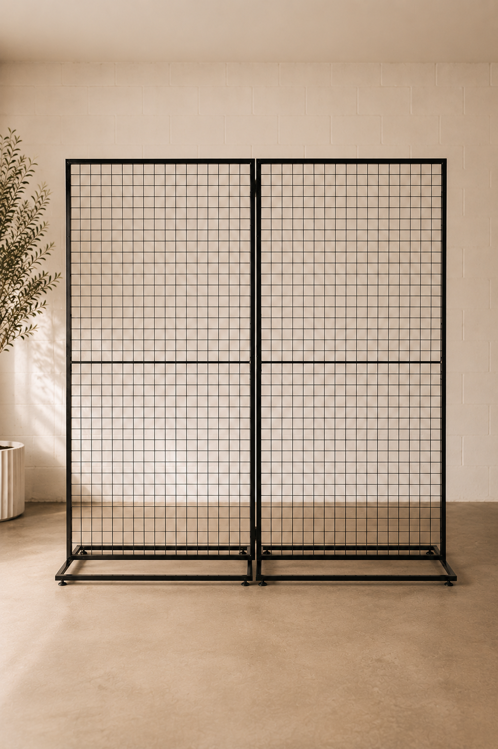 A black metal grid partition screen with two hinged panels, standing in a minimalist beige room with a concrete floor and a potted plant on the left.