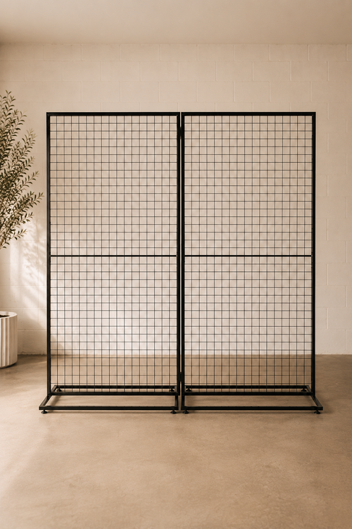 A black metal grid partition screen with two hinged panels, standing in a minimalist beige room with a concrete floor and a potted plant on the left.
